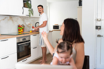 Happy family multitasking at home, father cooking in kitchen while young mother working on laptop and caring for cute toddler baby, balancing domestic life and remote work