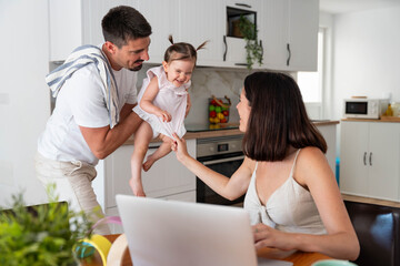 Happy family balancing work and parenthood, father playing with laughing child while mother uses laptop working from home in modern kitchen