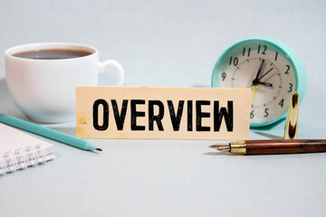 There is a cup of coffee on a wooden table, a clock, glasses and a notebook with the word OVERVIEW.