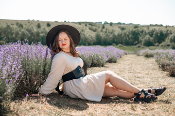 Woman sits in lavender field wearing a black hat and enjoying the sunlight during a warm day in summer