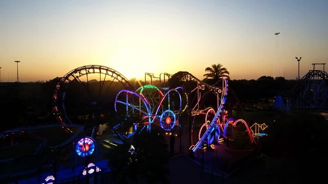 The amusement park features illuminated roller coasters and vibrant lights as the sun sets. The colorful lights of the amusement park create a lively atmosphere during twilight.