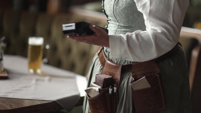Unrecognizable waitress in a dirndl entering an order on a mobile terminal, modern contactless payment in a tavern