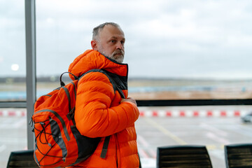 A man in an orange jacket and gray beard waits in the airport terminal