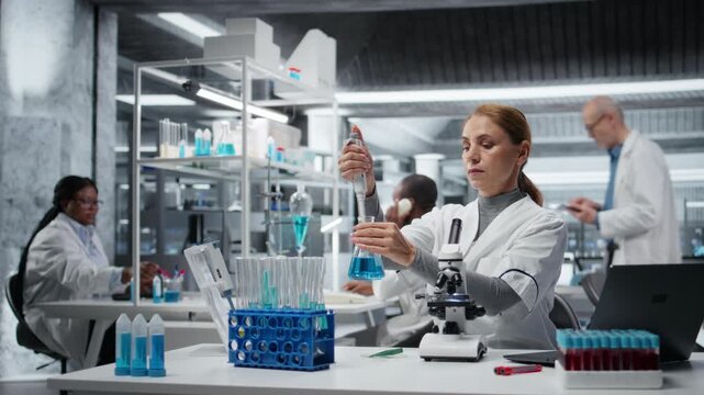 Female researcher in lab pipetting blue liquid into test tube to analyze it. Middle aged woman transferring chemical sample to prepare for immediate spectroscopic analysis, camera A