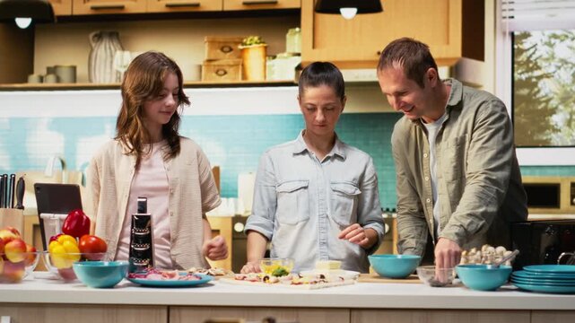 Tween daughter taking a family photo of her and parents during cooking time, smiling for a picture while they prepare homemade pizza in the kitchen. Fun weekend activity together. Camera B.