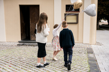 Children dressed formally walking on cobblestone pavement holding balloons near a historic building, candid outdoor moment capturing celebration, childhood, and timeless European atmosphere.
