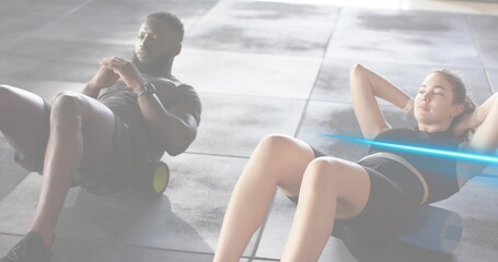 Rolling partners doing exercises on gym floor with foam roller and block, wearing black sportswear