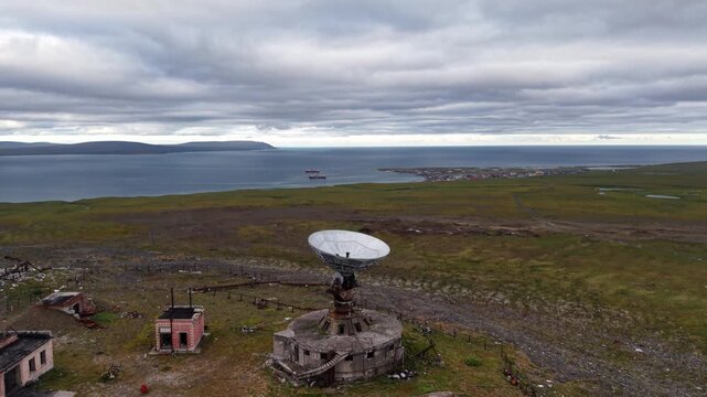 Aerial view of a parabolic dish at the Orbita space communications station near Lavrentiya. Broad tundra and coastline lie under moody sky, industrial detail and wild scenery deliver Arctic value