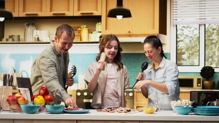 African American family enjoying silly karaoke session for pizza day in the kitchen, singing with utensils and doing goofy dance moves together. Weekend activity and cooking time. Camera B.