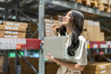 Professional asian female warehouse worker talking on walkie-talkie while holding laptop, logistics...