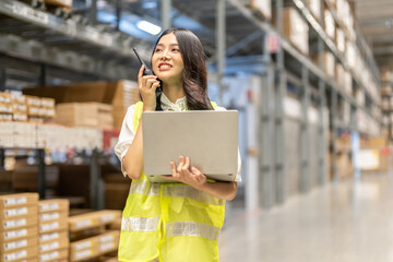 Fototapeta premium Professional asian female warehouse worker talking on walkie-talkie while holding laptop, logistics manager checking inventory in distribution center, supply chain and shipping business concept