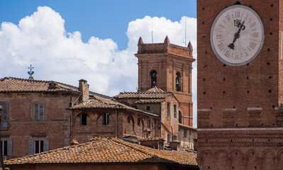 Naklejka premium Medieval clock tower and surrounding brick buildings in Siena, Italy. A classic example of Tuscan Gothic architecture under a bright blue sky.