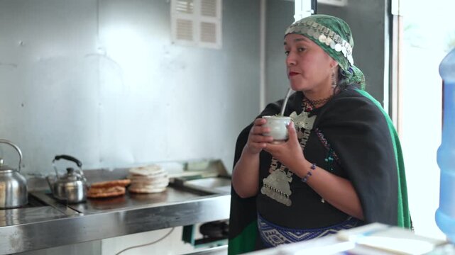 Mapuche woman in traditional clothing drinking yerba mate from a gourd in a kitchen