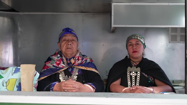 Two native mapuche women wearing traditional jewelry and clothing at their market stall