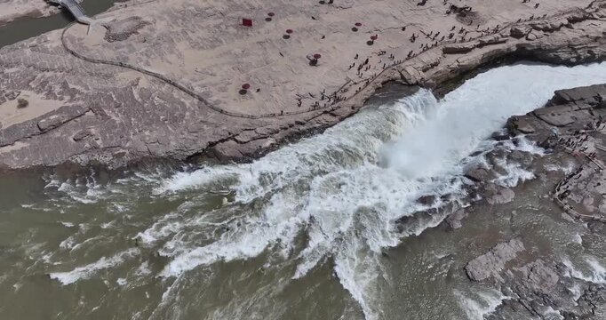 Aerial video of Hukou Waterfall, mighty Yellow River thunders through a dramatic gorge with tourists along viewing paths on the Shanxi-Shaanxi border, China.