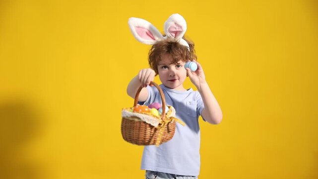 Easter celebration. Cute little boy in headband with bunny ears and wicker basket full of painted eggs on yellow background