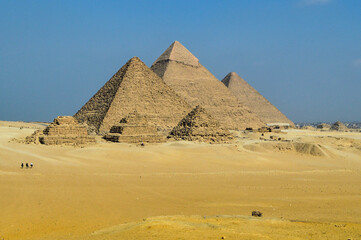 ​A scenic landscape of the Giza Necropolis featuring the three main Pyramids of Giza: Menkaure (left), Khafre (center), and Khufu (right). The foreground showcases the three smaller subsidiary Queens'