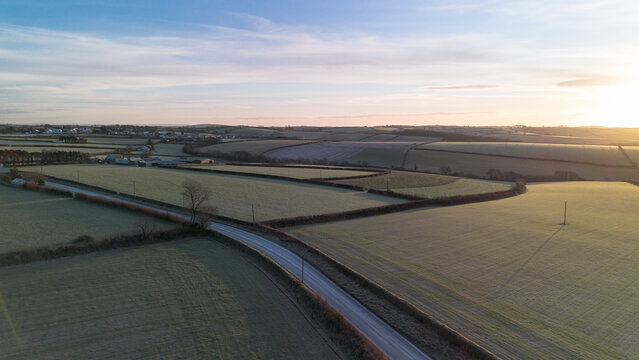 Aerial view of rural agricultural fields and a winding country road during a frosty winter sunrise in Cornwall, England. Light frost covers the farmland, creating soft textures and muted tones across 