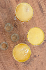 Overhead shot of three vibrant mango cocktails in glasses alongside empty shot glasses on a wooden surface. the bright yellow drinks contrast with the warm wood texture.