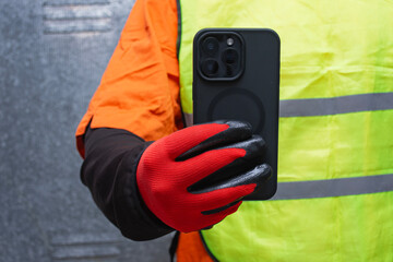 Construction worker wearing protective gloves and reflective safety vest while holding a smartphone. Concept of modern industrial communication, safety at work, and digital technology in construction.