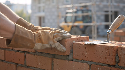 Construction worker hands in protective gloves laying red bricks on a wall. Masonry process with trowel and cement mortar on a building site