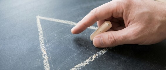 Close up of a hand holding white chalk drawing a geometric triangle shape on a textured dark surface or chalkboard with visible dust blackboard writing education human writing instrument