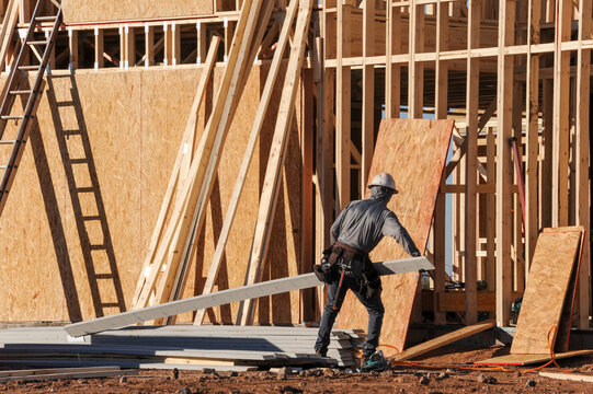 Construction worker carrying building materials at a wood framed residential structure under construction, representing housing development, structural assembly, and active jobsite operations
