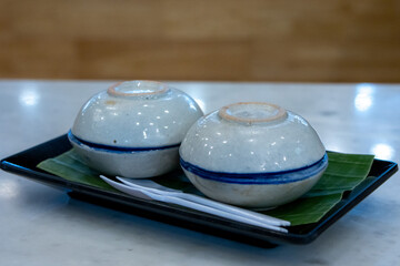 Kanom Tuay, Steamed Coconut Custard Two small ceramic bowls, flipped upside down, rest on a banana leaf on a black plate