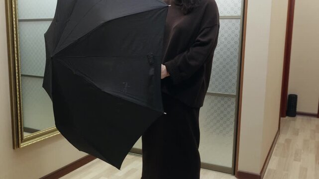 Adult woman holding black umbrella indoors, folding it closed near mirror. Preparation for weather, daily routine and home lifestyle concept.