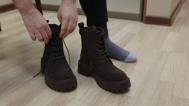 Adult woman bending indoors untangling dark boot shoelaces on laminate floor. Footwear preparation, patience and daily routine concept.