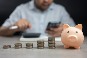 Coin like growing graph placed with piggy bank and blur background of Asian man calculating finances about future expenses or investments at home.