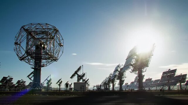 Array of Solar Panels and a Large Concentrator Dish Under a Bright Sun and Blue Sky in a Rural Landscape With Lens Flare Effect