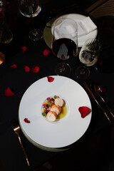 Elegant culinary presentation of a gourmet meal, showcasing a white plate with food, surrounded by wine glasses, flower petals and silverware