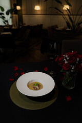 A culinary delight of soup, roses, and wine glasses, all set in a dimly lit restaurant, creating a romantic atmosphere. The dish is presented on a table, with rose petals scattered.
