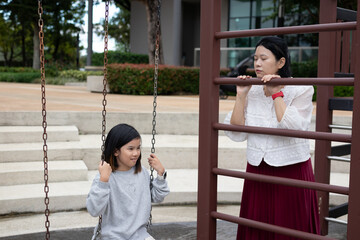 Happy Asian mother gently pushing her daughter on a swing in the outdoor playground, enjoying active family bonding time