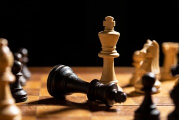 White chess king standing upright next to fallen black king on wooden board in darkness