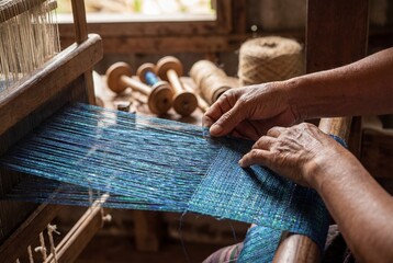 Close up of elderly hands weaving blue silk threads on a traditional wooden loom in a workshop