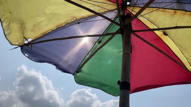 Sunlight filters through the colorful panels of an open umbrella against a bright blue sky with fluffy white clouds.