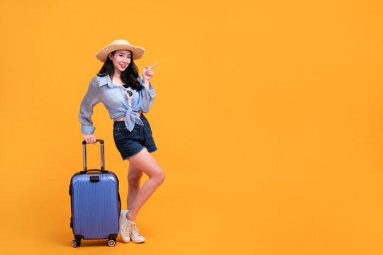 Happy Asian woman traveler standing and holding suitcase isolated on yellow background, Tourist girl having cheerful holiday trip concept, Full body composition.
