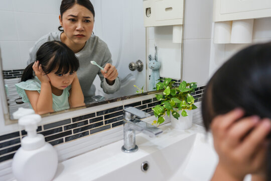 stubborn child girl refusing to brush her teeth in the bathroom. mother is trying to encourage her unhappy daughter during the morning routine, while child looks grumpy and resistant.