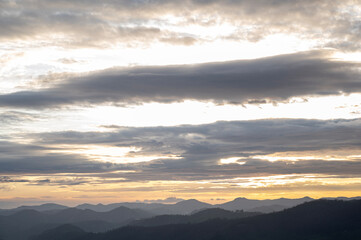 Obraz premium Landscape sunrise near Ooty, Nilgiris showing layered mountain silhouettes emerging through mist on a monsoon morning, with a cloudy sky glowing in soft black and yellow gradients.