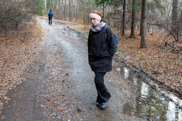 Fototapeta premium Upset caucasian girl in a black coat and scarf walking on a muddy forest path during autumn. Moody weather and cold season concept for outdoor activities.