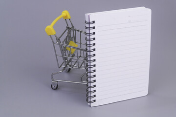 Shopping cart with blank notebook page on grey background. List of goods to buy concept.	