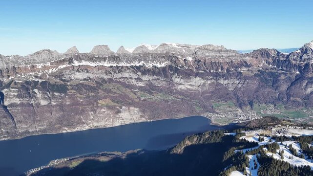 A wide aerial view of the Churfirsten limestone range above Walensee near Flumserberg, Switzerland, showing snow-covered slopes and dramatic alpine peaks under clear skies.