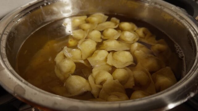Cappelletti cook slowly in a clear broth, floating inside a metal pot on the stove in a traditional kitchen.