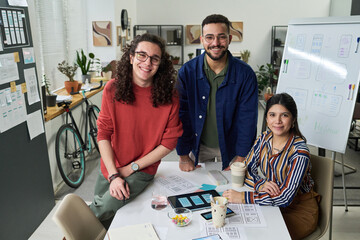Group portrait of three young adults, including two men and woman, collaborating on user interface design project, smiling and looking at camera in modern office