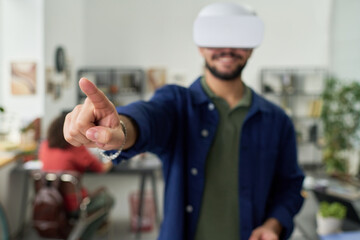 Young adult man wearing virtual reality headset interacting with digital interface, standing in modern workspace with outstretched arm and extended finger