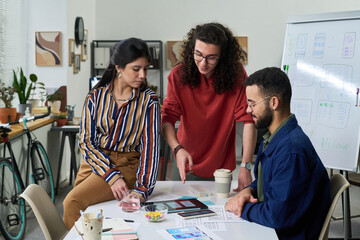 Multiethnic group of young adults collaborating on project, discussing digital interface designs at table with documents and tablet, creative workspace with brainstorming session