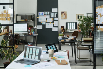 Modern office workspace showing digital devices on desk with open laptops, tablet, smartphone, documents, coffee cup, plants, and creative project boards in background