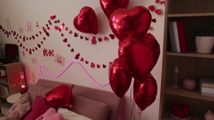 No people shot of bed with pink cushions, heart-shaped balloons, red paper chains, cards and neon lighting on wall, wooden bookcase in cozy bedroom prepared for Valentines day celebration